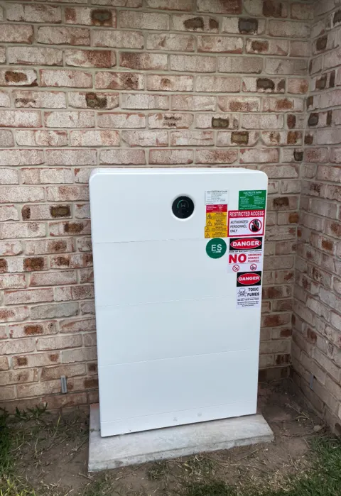 Home battery with protective cover fitted — outdoor install against brick wall